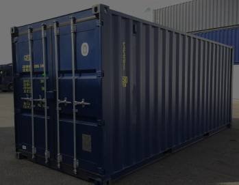 Новый синий 20-футовый морской контейнер стоит на бетонной площадке в солнечный день, видна маркировка и желтые полосы на углах, на заднем плане складские постройки и голубое небо с легкими облаками