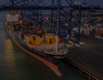 Ночной вид грузового судна, загруженного контейнерами, в порту с кранами и контейнерными терминалами на фоне вечернего неба.