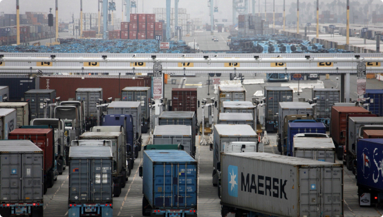 Длинная очередь грузовиков с контейнерами ждет въезда в морской порт в пасмурную погоду, виден контейнер Maersk на переднем плане, вдали портовые краны и штабели контейнеров