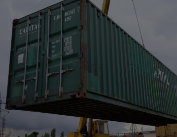 Зеленый 40-футовый морской контейнер с надписью 'CAPITAL' поднимается краном на фоне облачного неба и складской территории с другим контейнерами и линией электропередач