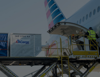 Желтый грузовик с белым фургоном припаркован у разгрузочной рампы склада с открытыми воротами, рядом стоит синий грузовик-рефрижератор, на заднем плане складское здание и голубое небо с облаками