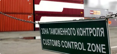 Зеленый знак 'ЗОНА ТАМОЖЕННОГО КОНТРОЛЯ / CUSTOMS CONTROL ZONE', закрепленный на цепи, на фоне сложенных морских контейнеров разных цветов