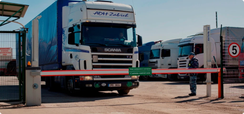Белый грузовик Scania с синим тентом и надписью пересекает таможенный пункт, охраняемый сотрудником в форме