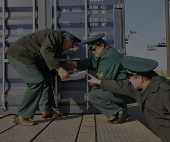 Три сотрудника таможни в форме осматривают запорное устройство морского контейнера, стоящего на железнодорожной платформе, сверяя данные с документами